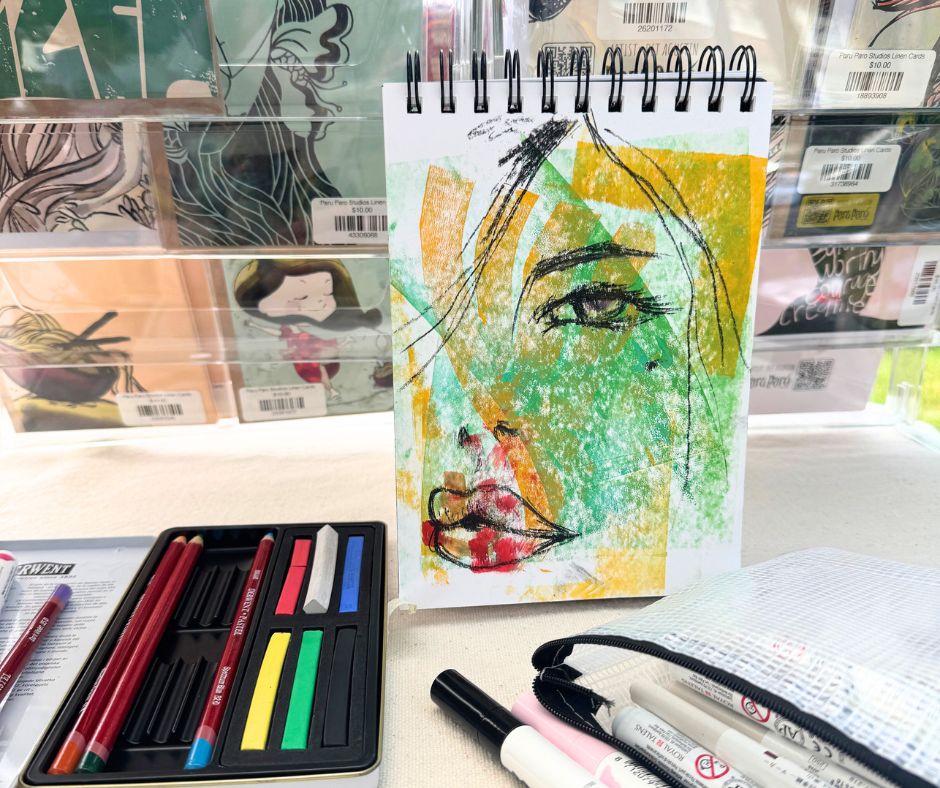 Hand-drawn portrait in a spiral sketchbook with layered green and yellow abstract background, surrounded by colorful art supplies and personalized journals at Sterling Street Market.